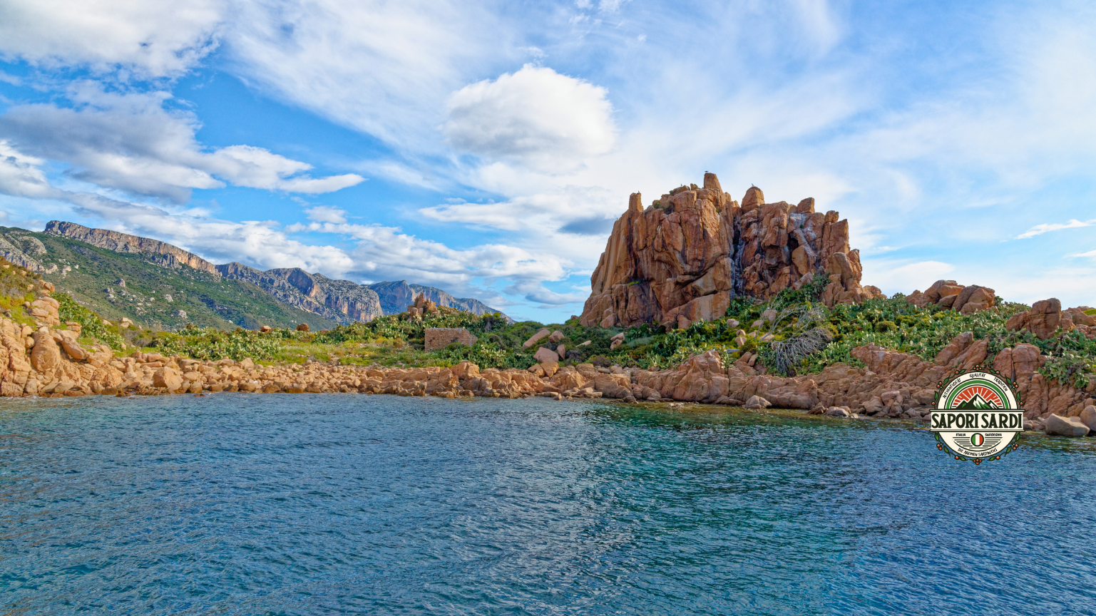 Vom Wasser aus ist Ogliastra auf Sardinien zu erkennen. Vorne an der üste ein großer Felsen und im Hinterland ein Bergzug. Die Landschaft ist saftig und grün sowie von Felsen durchzugen. Unten rechts im Bild ist das Logo von Sapori Sardi zu erkennen.