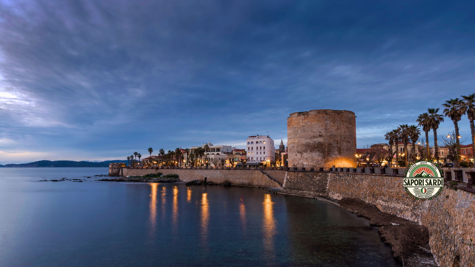Ein Bild vom Torre Sulis Alghero auf Sardinien. Es ist Abend und der Himmel ist dunkelblau ebenso wie das Meer in welchem sich die Lichter der Promenade spiegeln. Entlang der Promenade stehen Palmen entlang der alten Steinmauer. Unten rechts im Bild ist das Logo von Sapori Sardi.